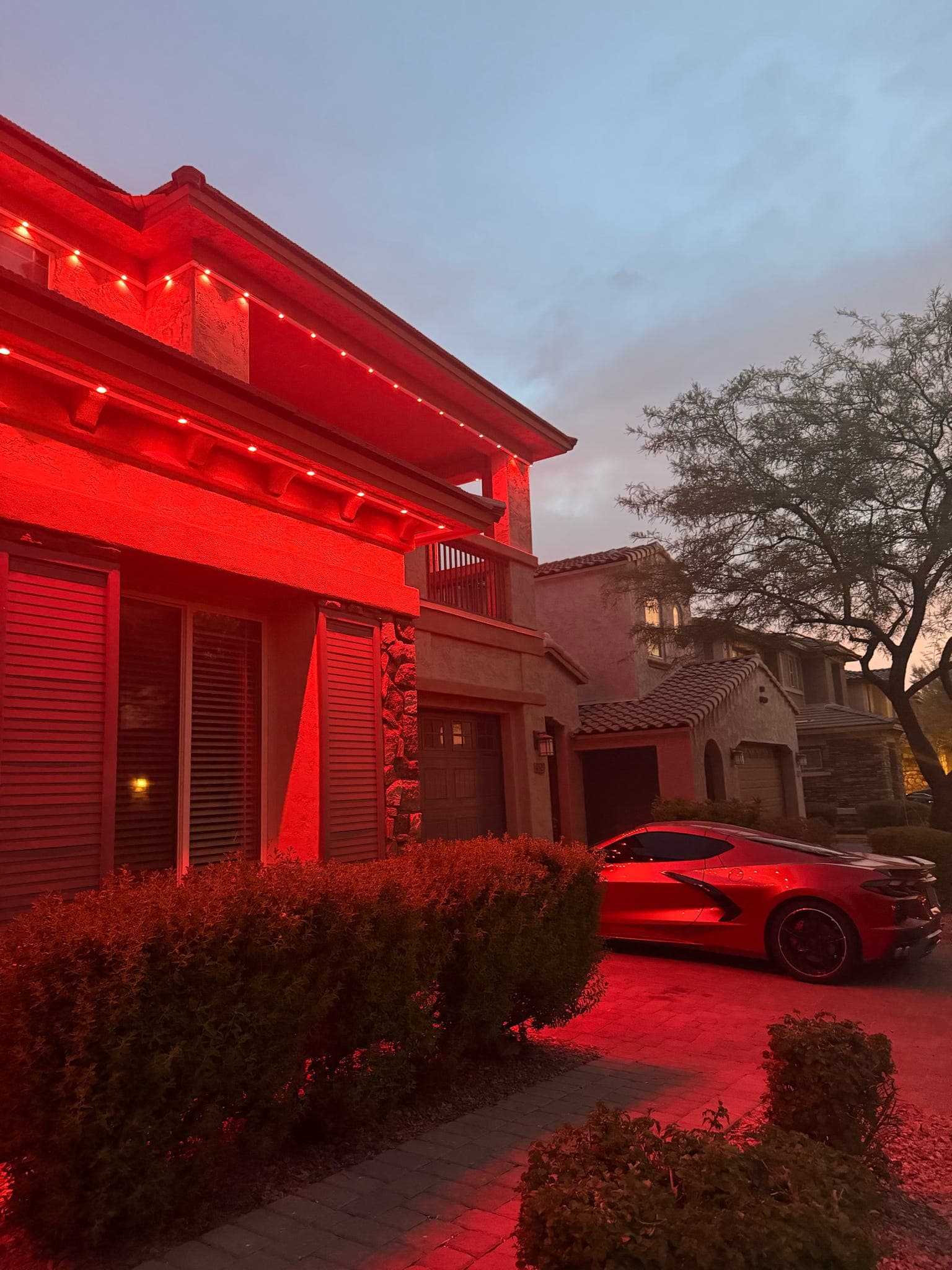 Side view of the same Phoenix two-story stucco home showing the second-story balcony overhang lit in red by Govee Permanent Outdoor Pro pucks mounted in GeauxTrax PETG track